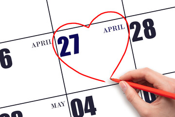A woman's hand drawing a red heart shape on the calendar date of 27 April. Heart as a symbol of...