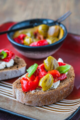 Open bruschetta sandwich with curd cheese, marinated roasted peppers and grilled olives on a red ceramic plate on a wooden background. Sandwich recipes. Antipasti.