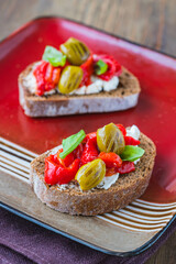 Open bruschetta sandwich with curd cheese, marinated roasted peppers and grilled olives on a red ceramic plate on a wooden background. Sandwich recipes. Antipasti.