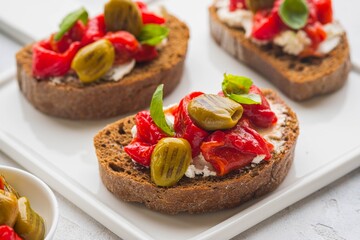 Open bruschetta sandwich with curd cheese, marinated roasted peppers and grilled olives on a white ceramic board on a light background. Sandwich recipes. Antipasti.