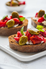 Open bruschetta sandwich with curd cheese, marinated roasted peppers and grilled olives on a white ceramic board on a light background. Sandwich recipes. Antipasti.