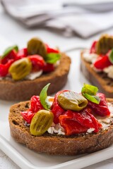 Open bruschetta sandwich with curd cheese, marinated roasted peppers and grilled olives on a white ceramic board on a light background. Sandwich recipes. Antipasti.