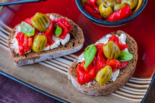 Open bruschetta sandwich with curd cheese, marinated roasted peppers and grilled olives on a red ceramic plate on a wooden background. Sandwich recipes. Antipasti.
