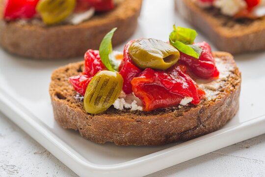 Open bruschetta sandwich with curd cheese, marinated roasted peppers and grilled olives on a white ceramic board on a light background. Sandwich recipes. Antipasti.