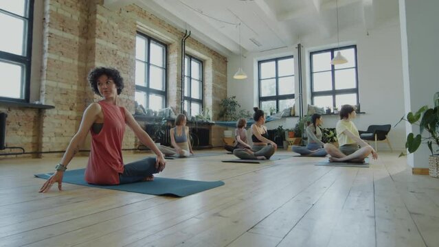 Zoom in shot of group of women in sportswear practicing seated spinal twist on mats during yoga class with female instructor