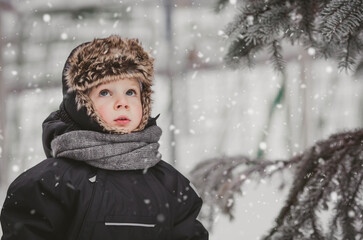 Fototapeta na wymiar Portrait of a beautiful little European boy in winter under a snow-covered fir tree in the park. The concept of childhood and lifestyle.