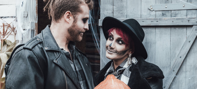 Scary love family couple man,woman celebrating halloween with pumpkin jack-o-lantern.Terrifying black skull half-face makeup,witch costumes,stylish images,jacket,hat. Fun,holiday party at street barn