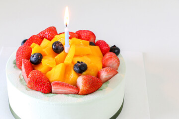 Fruit birthday cake with fresh strawberries, blueberries and mango, and light blue candle lit with flame, on left of frame