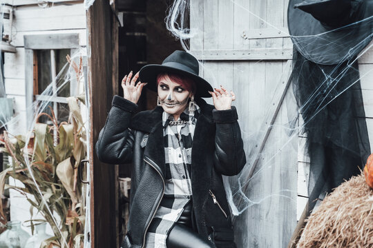 Scary woman, girl celebrating halloween.Terrifying black skull half-face skeleton makeup and witch costume in stylish image. Jacket, hat.Horror,fun at photoshoot, holiday party near barn on street