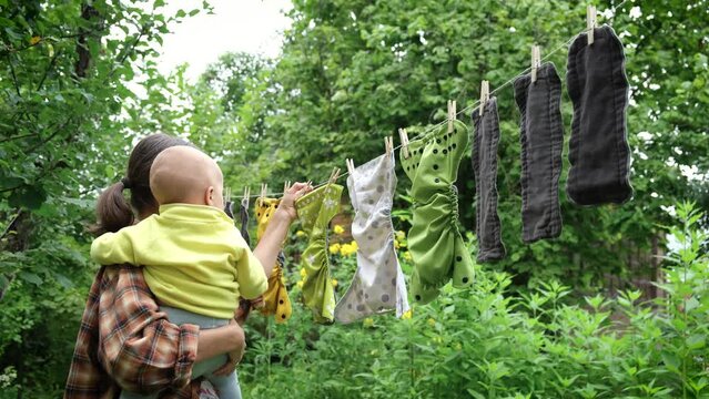 Mother with her baby removes reusable diapers drying on clothesline in garden. Modern eco friendly cloth nappy for infant child hygiene. Zero waste concept.