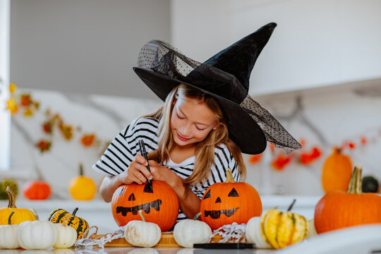 Girl in witch hat drawing faces on pumpkins for halloween on white kitchen.