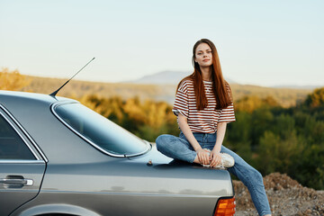 A woman car driver sits on the trunk of a car and looks into the distance admiring a beautiful view...