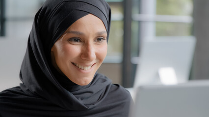 Cute positive arab woman in hijab looks at laptop screen smiling watching funny video on internet reads good news happy female professional manager working on computer in office gets nice notification