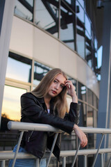 Fototapeta na wymiar a blonde girl in a leather jacket and light jeans stands near modern building and looking into camera