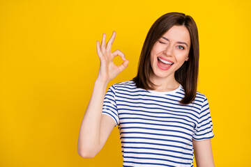 Photo portrait of cheerful young lady show okey sign winking quality approval wear stylish striped look isolated on yellow color background