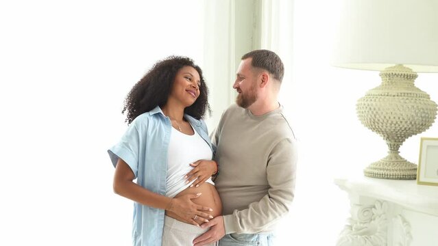 White man touching belly of his pregnant black wife. Pregnant woman and loving husband hugging tummy at home. Feeling baby movement by multiethnic couple on pregnant belly.