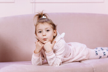 Fototapeta premium Little cute adorable Caucasian girl is lying alone on pink sofa and looking in camera in room. High quality photo