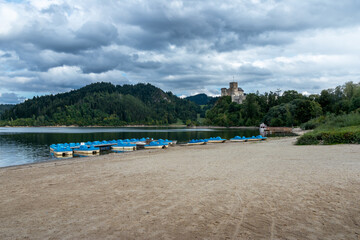 Fototapeta na wymiar Medieval castle in Niedzica, Poland seen from the beach of the Czorsztynskie Lake. Blue pedal boats moored at the sandy shore. Pieniny mountains in the horizon. Cloudy, summer day.