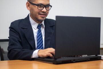 A happy man wearing glasses and a tie smiles while he works on his laptop.