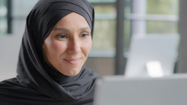 Cute positive arab woman in hijab looks at laptop screen smiling watching funny video on internet reads good news happy female professional manager working on computer in office gets nice notification