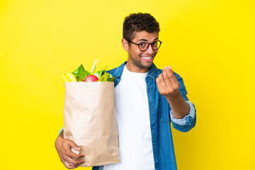 Young Brazilian man holding a grocery shopping bag isolated on yellow background making money gesture