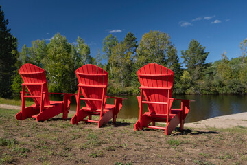 Red wooden Muskoka Adirondack chairs by a pier in front of the lake, beautiful sunny day, blue sky...