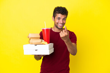 Young brazilian man holding pizzas and burgers isolated background doing coming gesture