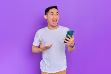 Shocked young Asian man in white t-shirt using smartphone and looking away isolated on purple background