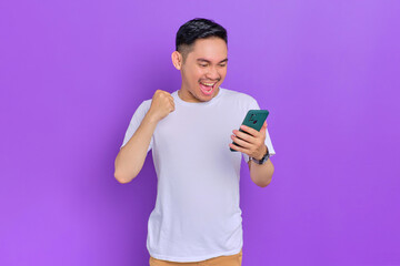 Excited young Asian man in white t-shirt using smartphone and making winner gesture isolated on purple background