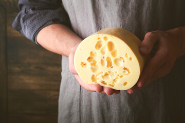 Hands of man hold two big slices of cheese maasdam against background of old wooden wall in cheese factory. Cheesemaker hold cheese maasdam in hand. Free space for your text.