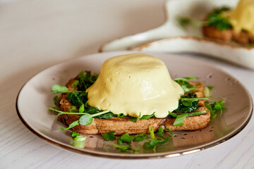 Close up of creative vegan alternative eggs benedict dish with sourdough bread, creamy plant-based hollandaise sauce, tofu, young onion, greens, pea sprouts.