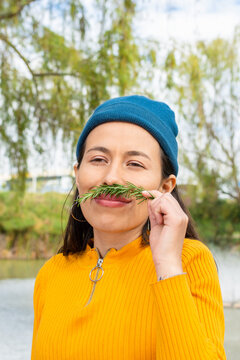 Latin Young Woman In A Blue Beanie And Yellow Sweater Outdoors Holding A Branch Of Rosemary And Enjoying Its Smell