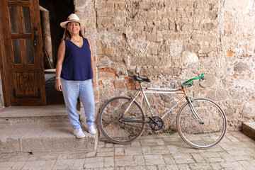 Latina morena parada en la puerta junto a una bicicleta - obrazy, fototapety, plakaty