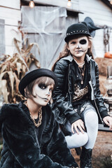 Beautiful scary little girls celebrating halloween. Terrifying black, white half-face makeup,witch costume, stylish image. Horror, fun at children's party in barn on street. Hat, jacket