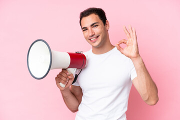 Young caucasian man isolated on pink background holding a megaphone and showing ok sign with fingers