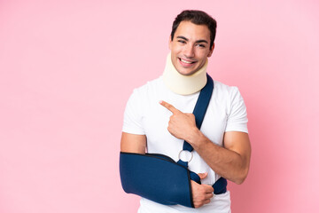 Young caucasian man with broken arm and wearing a sling isolated on pink background pointing to the...