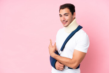 Young caucasian man with broken arm and wearing a sling isolated on pink background pointing back