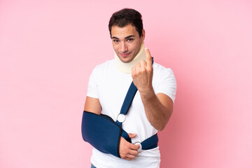 Young caucasian man with broken arm and wearing a sling isolated on pink background doing coming...