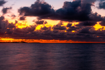 Fototapeta premium Dramatic sunset over calm ocean waters, vibrant orange and purple clouds reflecting on the sea, silhouetted distant shoreline and tranquil horizon.