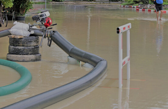 Pipes lead to a pump working on a flooded residential street. A flood barrier warns that the machine is in operation.  Blurred in the background a person can be partially seen walking in the water.