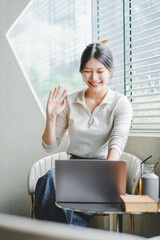 Attractive and happy young female Asian student studying online, sitting at desk, using laptop computer, having video chat, waving, education concept.