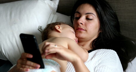 Mother using cellphone device at home lying in bed with sleeping baby on chest napping
