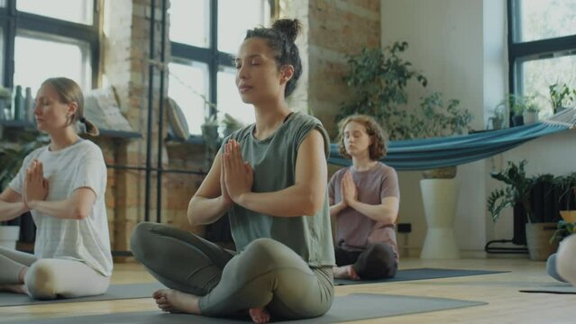Beautiful biracial woman sitting in lotus pose with eyes closed and holding hands in Namaste and mudras during group yoga class