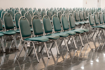 Many empty green leather chairs in a wide hall room, lights on. A floor reflect the objects. Give a feeling of emptiness.