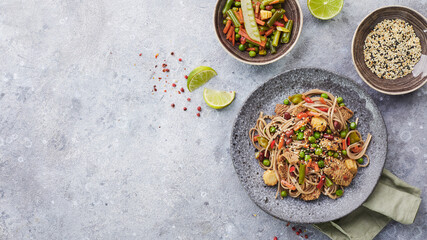 Wok with turkey meat, soba noodles, corn, green peas, green beans and carrots served on gray background with chopsticks. Asian food, concept of street food