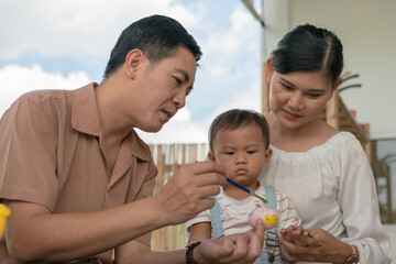 Obraz premium Asian family with baby little son painting eggs with brush in class workshop. Happy Asian father and mother with little son and teaching painting eggs with water colour