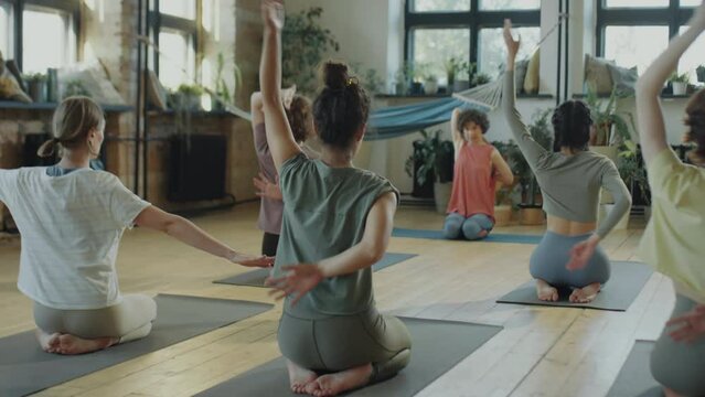 Group of women sitting on mats and bringing hands together behind back while doing cow face pose during yoga practice with female teacher in studio