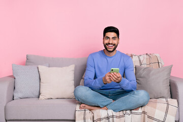 Photo of cute cheerful arab guy dressed blue pullover sitting couch chatting modern device empty space isolated pink color background
