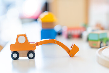 An orange wooden toy excavator is placed on the table in front of other blurry wooden toys.