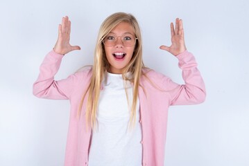 caucasian blonde little girl wearing pink jacket and glasses over white background looks with excitement at camera, keeps hands raised over head, notices something unexpected, reacts on sudden news.